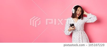 Carefree asian woman having fun, dancing and listening music in headphones, holding mobile phone, standing in dress over pink background 105107220