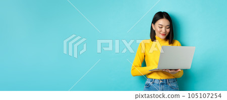 Beautiful asian female student working on laptop, typing on keyboard and looking at screen with pleased smile, standing over blue background 105107254