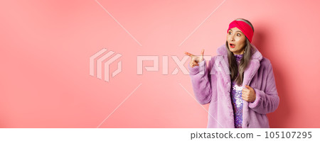 Fashion and shopping concept. Trendy elderly asian female in fake fur and glittering dress pointing, looking left with amazement, checking out offer, pink background 105107295