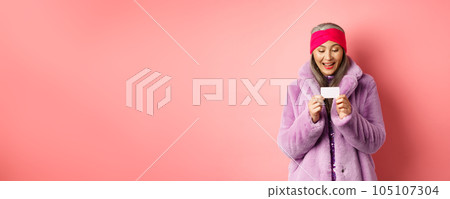 Shopping and fashion concept. Beautiful and stylish asian woman looking at plastic credit card, smiling pleased, standing over pink background Shopping and fashion concept. Beautiful and stylish asian woman looking at plastic credit card, smiling pleased, standing over pink background 105107304
