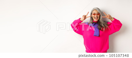 Fashion and beauty concept. Image of stylish asian senior woman in sunglasses smiling, showing peace signs and looking happy, standing over white background Fashion and beauty concept. Image of stylish asian senior woman in sunglasses smiling, showing peace signs and looking happy, standing over white background 105107348
