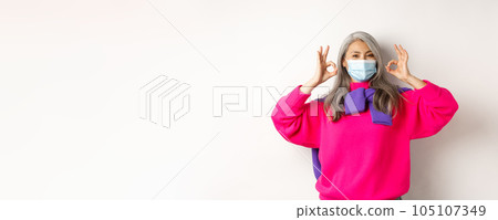 Covid-19, quarantine and lockdown concept. Fashionable asian old woman recommending to wear medical mask, showing okay signs in approval, standing over white background Covid-19, quarantine and lockdown concept. Fashionable asian old woman recommending to wear medical mask, showing okay signs in approval, standing over white background 105107349