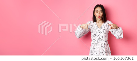 Impressed asian woman in dress pointing fingers down, look there gesture, check out discounts and sales, standing amazed over pink background Impressed asian woman in dress pointing fingers down, look there gesture, check out discounts and sales, standing amazed over pink background 105107361