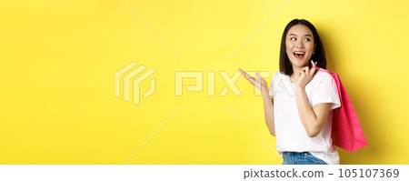 Cheerful asian female shopper looking amused, holding shopping bag and looking right, standing over yellow background Cheerful asian female shopper looking amused, holding shopping bag and looking right, standing over yellow background 105107369