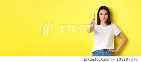Confident and serious woman tell no, showing extended finger to stop and prohibit something bad, frowning and looking at camera self-assured, standing over yellow background 105107370