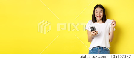 E-commerce and online shopping concept. Cheerful asian girl paying in internet, holding smartphone and plastic credit card, smiling and looking left, yellow background 105107387
