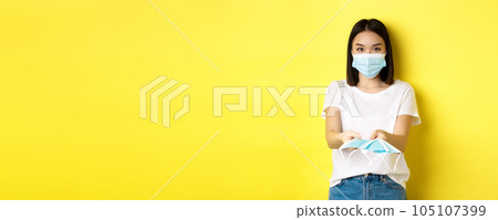 Coronavirus, quarantine and medicine concept. Cute asian girl giving medical masks and looking at camera, yellow background Coronavirus, quarantine and medicine concept. Cute asian girl giving medical masks and looking at camera, yellow background 105107399