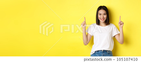 Beauty and fashion concept. Beautiful asian woman in white t-shirt pointing fingers up, standing over yellow background 105107410