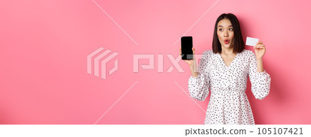 Cute asian woman shopping online, showing bank credit card and mobile screen, smiling and looking left at copy space, standing over pink background Cute asian woman shopping online, showing bank credit card and mobile screen, smiling and looking left at copy space, standing over pink background 105107421