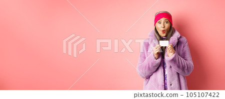 Shopping and fashion concept. Image of amazed asian senior woman checking out promo offer, holding plastic credit card and stare impressed at camera, pink background 105107422
