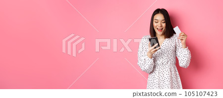 Attractive young asian woman order online, holding credit card and mobile phone, making internet purchase, standing happy over pink background Attractive young asian woman order online, holding credit card and mobile phone, making internet purchase, standing happy over pink background 105107423