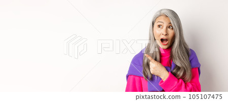 Close-up of amazed and intrigued asian senior woman pointing finger left at something cool, staring at camera promo offer, standing over white background Close-up of amazed and intrigued asian senior woman pointing finger left at something cool, staring at camera promo offer, standing over white background 105107475