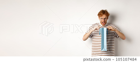 Surprised boyfriend open shopping bag with valentines day gift, staring inside with amazed face, standing over white background 105107484