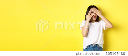 Intrigued asian girl waiting for surprise, peeking through fingers on eyes, smiling happy, standing over yellow background Intrigued asian girl waiting for surprise, peeking through fingers on eyes, smiling happy, standing over yellow background 105107488