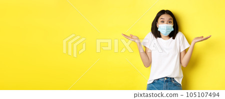Covid-19, quarantine and social distancing concept. Clueless asian woman in medical mask shrugging shoulders, spread hands sideways clueless, know nothing, yellow background 105107494