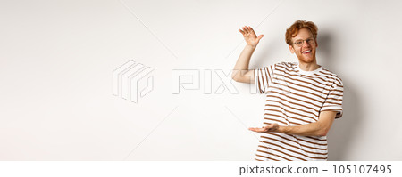 Happy and proud redhead guy with glasses showing something big, shaping large object with hands over white background, smiling at camera 105107495