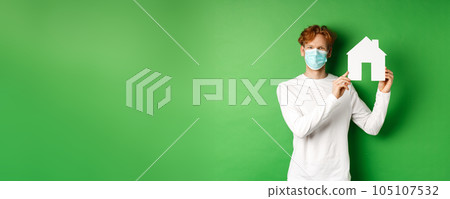 Real estate and covid-19 pandemic concept. Young redhead man in medical mask showing paper house cutout and smiling with eyes, standing over green background Real estate and covid-19 pandemic concept. Young redhead man in medical mask showing paper house cutout and smiling with eyes, standing over green background 105107532