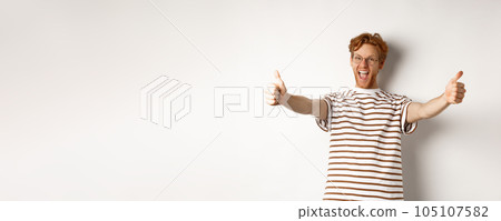 Happy redhead man scream yes and showing thumbs-up, approve and praise excellent company, standing over white background Happy redhead man scream yes and showing thumbs-up, approve and praise excellent company, standing over white background 105107582
