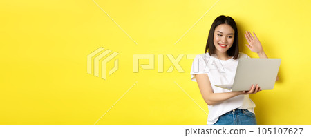 Happy asian woman video chat on laptop, waiving hand at computer camera and saying hello, standing over yellow background Happy asian woman video chat on laptop, waiving hand at computer camera and saying hello, standing over yellow background 105107627