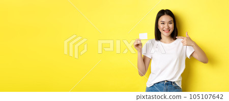 Young asian woman in casual white t-shirt showing plastic credit card and smiling at camera, yellow background Young asian woman in casual white t-shirt showing plastic credit card and smiling at camera, yellow background 105107642