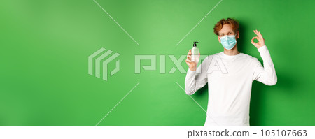 Covid-19, virus and social distancing concept. Smiling young man with red hair, wearing face mask from coronavirus, showing okay sign and hand sanitizer, green background 105107663