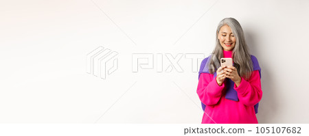 Beautiful asian senior woman in pink stylish sweater using smartphone. Old lady reading on mobile phone and smiling, standing over white background Beautiful asian senior woman in pink stylish sweater using smartphone. Old lady reading on mobile phone and smiling, standing over white background 105107682