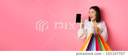 Attractive asian woman showing smartphone app and shopping bags, buying online via application, standing over pink background Attractive asian woman showing smartphone app and shopping bags, buying online via application, standing over pink background 105107707