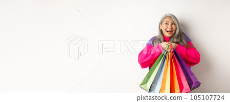 Fashionable senior asian woman shopaholic, hugging shopping bags and smiling joyful, buying with discounts, standing over white background Fashionable senior asian woman shopaholic, hugging shopping bags and smiling joyful, buying with discounts, standing over white background 105107724