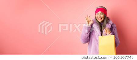 Shopping and fashion. Happy asian senior woman in stylish clothes holding paper bag from store, showing OK sign, recommending shop, pink background Shopping and fashion. Happy asian senior woman in stylish clothes holding paper bag from store, showing OK sign, recommending shop, pink background 105107743
