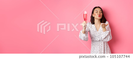 Romantic asian woman raising glass of champagne and sending air kiss at camera, celebrating and having fun, standing over pink background Romantic asian woman raising glass of champagne and sending air kiss at camera, celebrating and having fun, standing over pink background 105107744