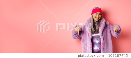 Fashion and shopping concept. Smiling senior asian woman pointing fingers down, showing store advertisement on bottom, wearing stylish purple winter coat, pink background Fashion and shopping concept. Smiling senior asian woman pointing fingers down, showing store advertisement on bottom, wearing stylish purple winter coat, pink background 105107745