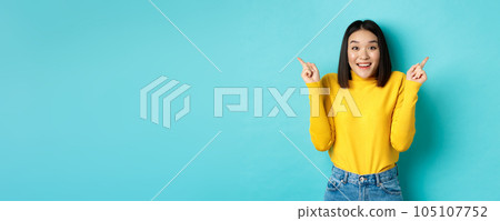 Beauty and fashion concept. Cheerful asian girl showing two promo offers, pointing fingers sideways at left and right advertisements and smiling, standing over blue background Beauty and fashion concept. Cheerful asian girl showing two promo offers, pointing fingers sideways at left and right advertisements and smiling, standing over blue background 105107752