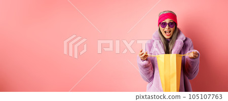 Shopping and fashion concept. Stylish asian elderly woman in sunglasses and faux fur coat open paper bag with gifts, looking amazed at camera, pink background Shopping and fashion concept. Stylish asian elderly woman in sunglasses and faux fur coat open paper bag with gifts, looking amazed at camera, pink background 105107763
