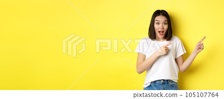 Beauty and fashion concept. Beautiful asian woman in white t-shirt pointing fingers right, demonstrate logo standing over yellow background Beauty and fashion concept. Beautiful asian woman in white t-shirt pointing fingers right, demonstrate logo standing over yellow background 105107764