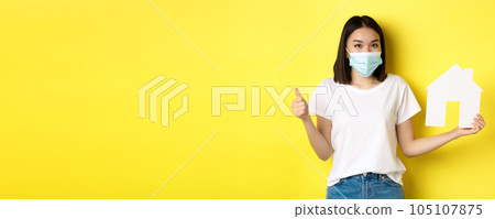 Covid-19, pandemic and real estate concept. Pretty asian woman smiling in medical mask, showing paper house cutout and thumb up, approve and like agency for buying property 105107875
