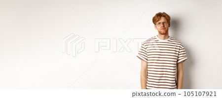 Reluctant and unamused redhead young man staring at camera, looking tired or displeased, standing over white background Reluctant and unamused redhead young man staring at camera, looking tired or displeased, standing over white background 105107921