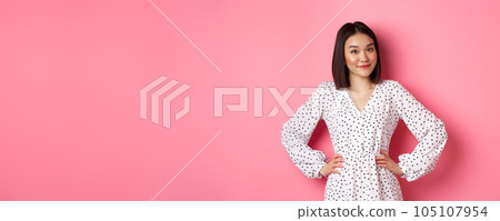 Beautiful korean lady in dress looking at camera and smiling, waiting for something, standing against pink background Beautiful korean lady in dress looking at camera and smiling, waiting for something, standing against pink background 105107954