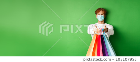 Happy young man in medical mask giving you shopping bags with purchases, smiling and wishing well, standing over green background. Covid-19 concept 105107955