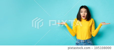 Confused and shocked asian woman dont understand, shrugging with spread hands, gasping and looking left, know nothing, standing over blue background 105108000