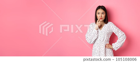 Troubled asian girl making decision, frowning and touching lip while thinking, looking unsure at upper left corner and choosing, standing over pink background 105108006