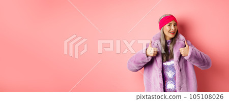 Fashion and shopping concept. Stylish asian granny saying yes. Trendy senior korean woman in winter fake-fur coat show thumbs-up in approval, praising good product, pink background Fashion and shopping concept. Stylish asian granny saying yes. Trendy senior korean woman in winter fake-fur coat show thumbs-up in approval, praising good product, pink background 105108026