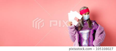 Covid-19 and real estate concept. Cool and stylish asian elderly woman in disco dress and medical mask showing paper house, standing over pink background Covid-19 and real estate concept. Cool and stylish asian elderly woman in disco dress and medical mask showing paper house, standing over pink background 105108027