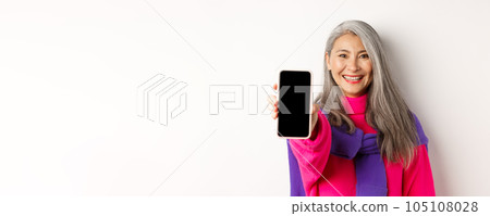 Online shopping. Close up of modern asian senior woman extending hand with mobile phone, showing blank smartphone screen and smiling, standing over white background 105108028