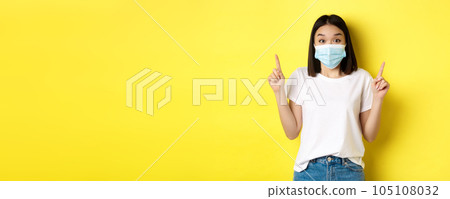 Covid-19, pandemic and social distancing concept. Young asian woman in white t-shirt and medical mask from coronavirus, pointing fingers up, showing special offer Covid-19, pandemic and social distancing concept. Young asian woman in white t-shirt and medical mask from coronavirus, pointing fingers up, showing special offer 105108032