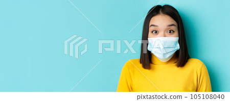 Covid-19, social distancing and pandemic concept. Close up of surprised asian woman in medical mask, raise eyebrows and look amazed at camera, blue background 105108040