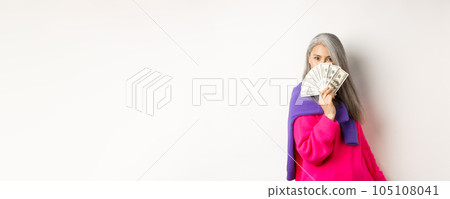 Shopping concept. Rich and fashionable korean senior woman hiding face behind money, looking at camera, standing over white background Shopping concept. Rich and fashionable korean senior woman hiding face behind money, looking at camera, standing over white background 105108041