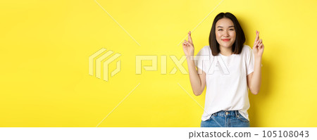 Hopeful asian girl smiling optimistic, feeling lucky, cross fingers and making wish, looking up dreamy, standing over yellow background Hopeful asian girl smiling optimistic, feeling lucky, cross fingers and making wish, looking up dreamy, standing over yellow background 105108043