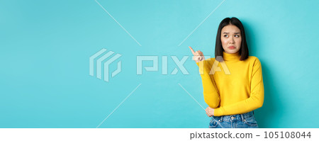 Shopping concept. Disappointed and gloomy asian woman sulking upset, pointing finger left at bad news banner, standing over blue background Shopping concept. Disappointed and gloomy asian woman sulking upset, pointing finger left at bad news banner, standing over blue background 105108044