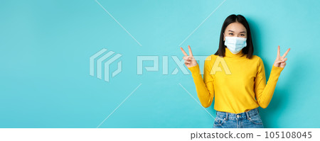 Covid-19, social distancing and pandemic concept. Kawaii asian woman showing peace signs and smiling happy, wearing medical mask from coronavirus disease, standing over blue background Covid-19, social distancing and pandemic concept. Kawaii asian woman showing peace signs and smiling happy, wearing medical mask from coronavirus disease, standing over blue background 105108045