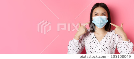 Covid-19, pandemic and lifestyle concept. Kawaii asian girl pointing fingers at her face mask, wearing preventive measures from coronavirus, smiling with eyes, pink background Covid-19, pandemic and lifestyle concept. Kawaii asian girl pointing fingers at her face mask, wearing preventive measures from coronavirus, smiling with eyes, pink background 105108049
